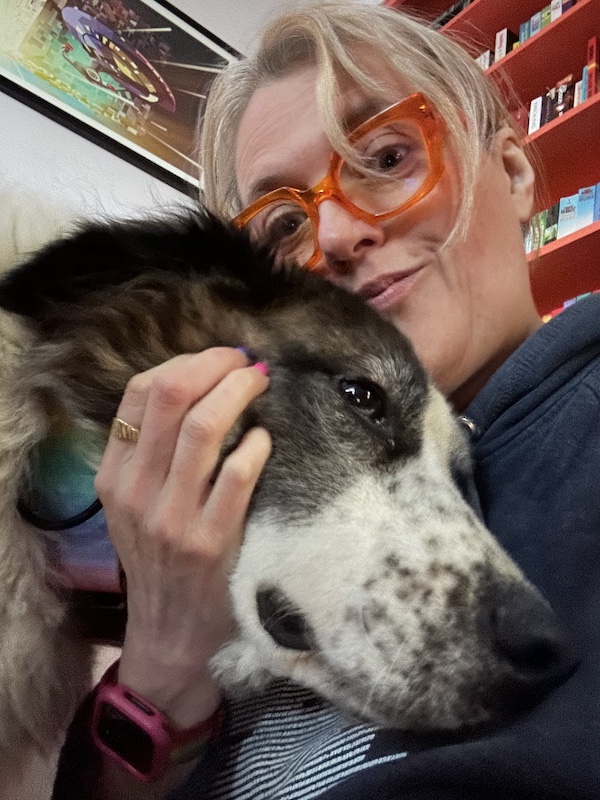 A brown and white fluffy dog snuggles into a human with blond hair and glasses. In the background there are books and an image of the starship Enterprise