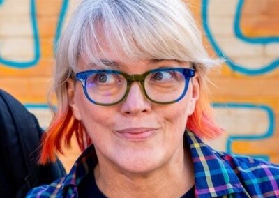 Portrait of the author with blond and sunset-coloured hair wearing glasses with blue-green frames. T-shirt says 'I came to make Conservatives uncomfortable'.